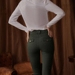 Person wearing a white long-sleeve shirt and green riding pants against a brown curtain background.
