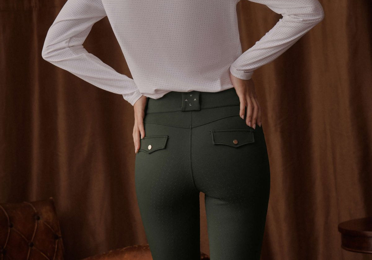 Person wearing a white long-sleeve shirt and green riding pants against a brown curtain background.
